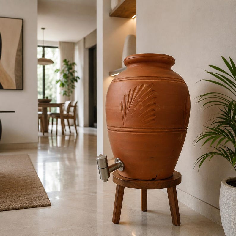 May include: A large, terracotta-colored ceramic water dispenser with a rounded body and a carved leaf pattern. It features a silver-toned metal spigot near the base and is supported by a small, round, three-legged wooden stand.