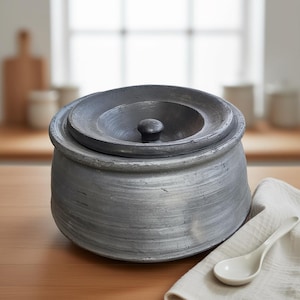 May include: A grey ceramic pot with a lid and a small knob. The pot has a textured surface and sits on a wooden table next to a white spoon and a white cloth.