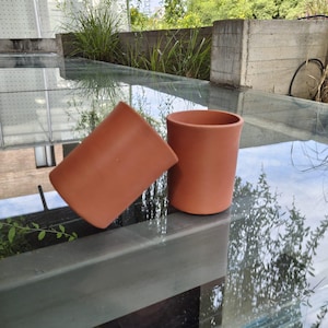 Könnte beinhalten: Zwei Keramikbecher in Terrakotta-Farbe. Ein Becher steht aufrecht, der andere ist auf die Seite gekippt. Die Becher haben eine glatte, unglasierte Oberfläche und eine einfache, zylindrische Form. Die Becher sind wahrscheinlich zum Trinken.