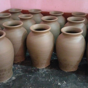 May include: A collection of multiple unglazed terracotta clay pots arranged in rows. Each pot features a rounded body, a wide neck, and a flared rim. The vessels have a smooth, matte finish with a consistent light brown color throughout the group.