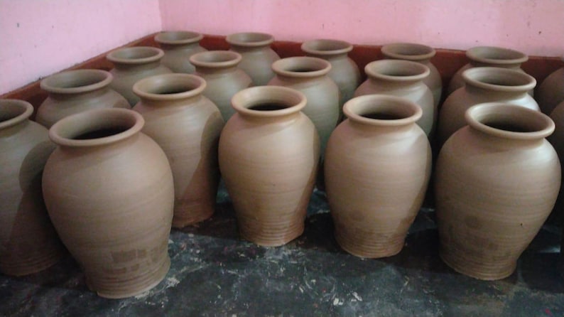 May include: A collection of multiple unglazed terracotta clay pots arranged in rows. Each pot features a rounded body, a wide neck, and a flared rim. The vessels have a smooth, matte finish with a consistent light brown color throughout the group.