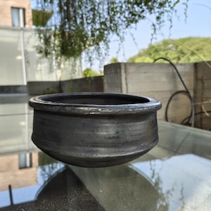 May include: A dark grey, handmade clay pot with a wide, rounded body and a slightly flared rim. The pot has a textured surface and appears unglazed. The pot sits on a glass surface, with a blurred outdoor background.