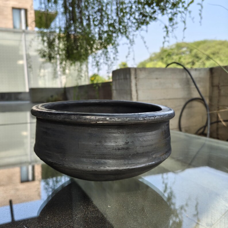 May include: A dark grey, handmade clay pot with a wide, rounded body and a slightly flared rim. The pot has a textured surface and appears unglazed. The pot sits on a glass surface, with a blurred outdoor background.