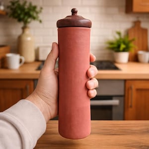 Może przedstawiać: Cylindryczny pojemnik w kolorze terakoty z ciemnobrązową drewnianą pokrywką. Pojemnik jest trzymany w dłoni, ukazując jego gładką fakturę i prosty design. Pokrywka ma mały, zaokrąglony uchwyt. Artykuły kuchenne.