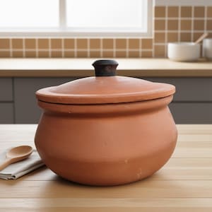 May include: A terracotta cooking pot with a lid and a black knob. The pot is round with a wide base and a slightly curved lid. It sits on a light-colored wooden surface. A wooden spoon and a folded cloth are visible on the left.