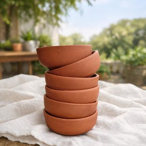 Może przedstawiać: Stos sześciu misek w kolorze terakoty. Miski są okrągłe i mają prosty design. Ułożone są na białej lnianej tkaninie. Tło jest rozmyte, ukazując zieleń i drewniany stół.