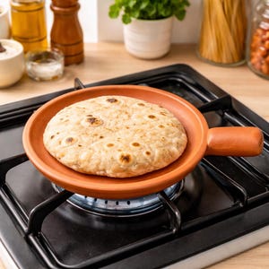 May include: A round, terracotta clay pan with a handle, holding a cooked flatbread. The pan sits on a black gas stove burner, with a blue flame visible. The flatbread is golden brown with darker spots.