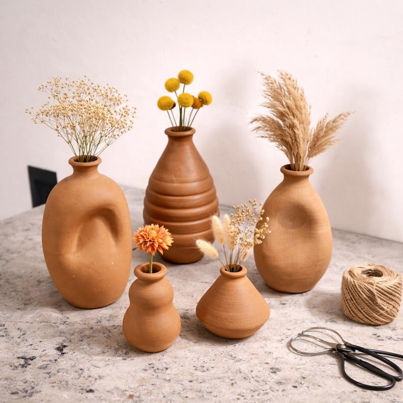 K&ouml;nnte beinhalten: Eine Sammlung von Terrakotta-Vasen in verschiedenen Formen, gef&uuml;llt mit getrockneten Blumen und Gr&auml;sern. Die Vasen variieren in ihrem Design, darunter eine gerippte Vase. Eine Schnurrolle und eine Schere sind ebenfalls vorhanden.