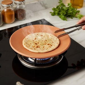 Puede incluir: Un plato de terracota con un pan plano cocido encima, humeando sobre un quemador de cocina de gas. Unas pinzas alcanzan el pan plano. Frascos de especias y hierbas frescas en el fondo.