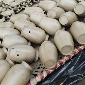 May include: A large collection of unglazed, light brown clay vessels and small circular pottery components. The vessels feature rounded bodies, narrow necks, and small circular openings on the sides, with some displaying subtle etched fan-like patterns.