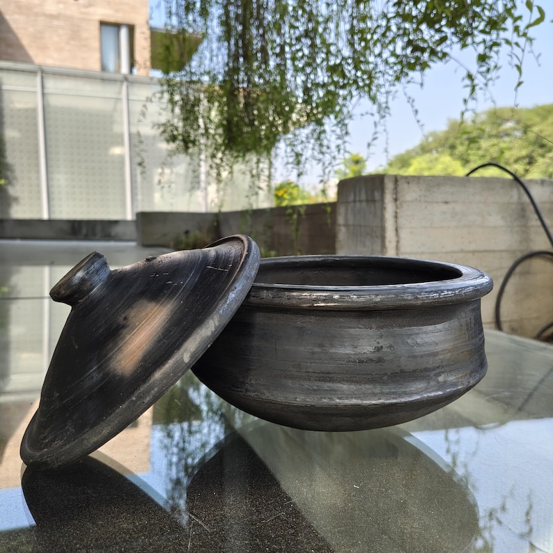 May include: A dark grey, handmade clay pot with a matching lid. The lid is slightly ajar, revealing the interior of the pot. The pot has a rounded shape with a wide opening, ideal for cooking and serving. The pot is on a reflective surface.