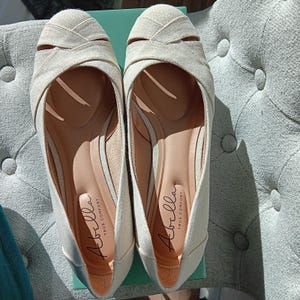 May include: A pair of beige Abella True Comfort flats. The shoes have a crisscross design on the top and a rounded toe. The interior is a light tan color with the Abella True Comfort logo. The shoes are on a light green box.