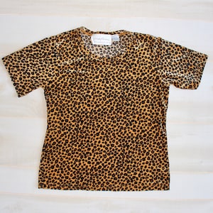 May include: A short-sleeved top with a leopard print pattern. The shirt features a scoop neckline and a fitted silhouette. The fabric is a blend of tan and black, creating a spotted design. The tag reads "Carolyn Taylor Collection".