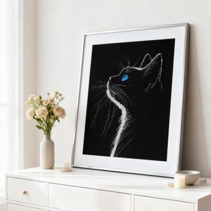May include: Framed black and white cat art print. The cat is depicted in profile, looking upwards with bright blue eyes. The artwork is in a silver frame and rests on a white dresser. A vase of flowers and a candle are also on the dresser.