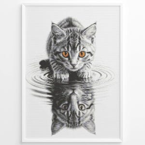 May include: Framed black and white print of a tabby cat with bright orange eyes, gazing at its reflection in rippling water. The cat's fur has a striped pattern. The print is in a white frame.