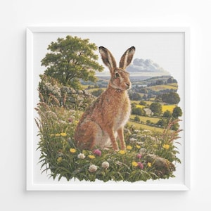 Könnte beinhalten: Eine quadratische Kreuzstichstickerei, die einen braunen Hasen in einem Feld mit Wildblumen zeigt. Der Hintergrund stellt eine hügelige Landschaft mit einem großen grünen Baum, Steinmauern und fernen Hügeln unter einem hellen Himmel dar.