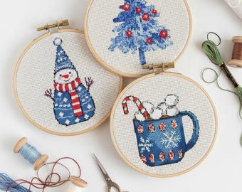 May include: Three cross-stitch hoops with holiday designs: a blue Christmas tree with red ornaments, a snowman in a blue hat and scarf, and a mug with candy canes and marshmallows. The hoops are set against a white background with sewing supplies.