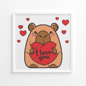 May include: A framed cross-stitch design featuring a cartoon bear holding a red heart with the words "I love you." The bear is light brown with a dark brown snout and ears. Small red hearts are scattered around the bear.