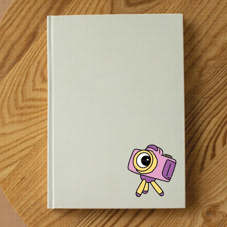 Pu&ograve; includere: Un taccuino bianco sporco con un motivo a griglia, con un adesivo di una fotocamera a cartoni animati rosa, giallo e viola. La fotocamera &egrave; su un treppiede. Il taccuino &egrave; su una superficie di legno.
