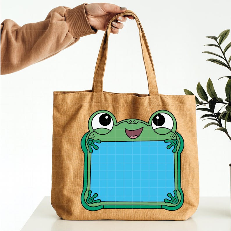 Pu&ograve; includere: Una borsa tote in tela beige con un grande ranocchio verde dei cartoni animati che tiene una griglia blu. La rana ha grandi occhi e una bocca sorridente. La borsa ha due manici.