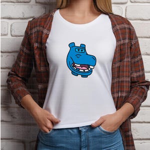 Pu&ograve; includere: T-shirt bianca con un disegno a cartoni animati di un ippopotamo blu. L'ippopotamo ha un corpo azzurro, una bocca rosa e denti bianchi. La persona indossa una camicia a quadri marrone e rossa.
