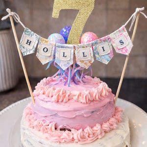 May include: A pink frosted cake with a banner that spells "HOLLIS" and a gold glitter "7" cake topper. The cake is decorated with pink frosting swirls and sits on a white plate. Colorful decorative balls are also on top.
