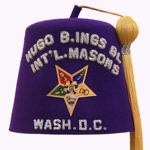 May include: A purple fez hat with the text "HUGO B.INGS BL INT'L.MASONS" and "WASH. D.C." in white, embellished with rhinestones. The hat features a gold tassel and a star emblem with various symbols.