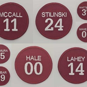 Peut inclure: Plusieurs écussons circulaires bordeaux avec des textes et des chiffres brodés en blanc. Les écussons affichent des noms tels que "MCCALL" et "STILINSKI" avec des numéros correspondants comme "11" et "24". D'autres écussons incluent "WHITTEMORE 37" et "MAHEALANI 06".