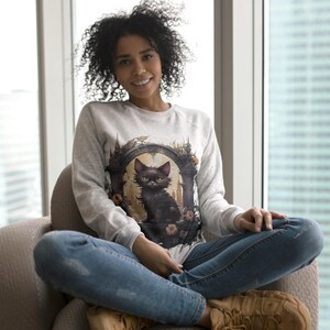 May include: A light gray sweatshirt featuring a black cat illustration within an arched frame. The cat has yellow eyes and is surrounded by floral accents. The sweatshirt is worn with blue jeans and brown boots.