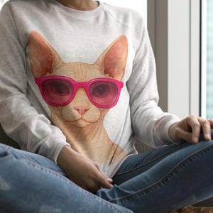 May include: A light gray sweatshirt featuring a graphic of a Sphynx cat wearing pink sunglasses. The cat's face is detailed with realistic fur and facial features. The sweatshirt is a casual style.