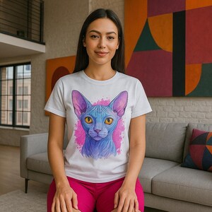 May include: White t-shirt featuring a blue cat illustration with pink accents. The cat has large ears and yellow eyes. The person is wearing pink shorts. The background includes a sofa and abstract art.