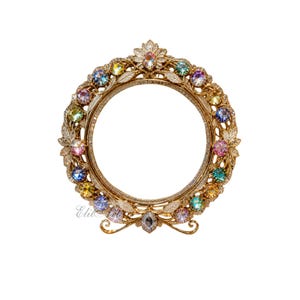 May include: A circular gold-toned metal picture frame featuring an ornate filigree design with leaf motifs. The frame is embellished with a series of round, multi-colored iridescent crystals and smaller clear stones. The text Elit Art is visible.