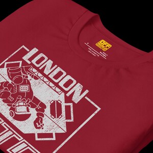 May include: A dark red crew neck t-shirt featuring a white distressed graphic print. The design includes the word LONDON in bold capital letters above a stylized illustration of a figure and a geometric shape. A yellow brand label is inside the neck.