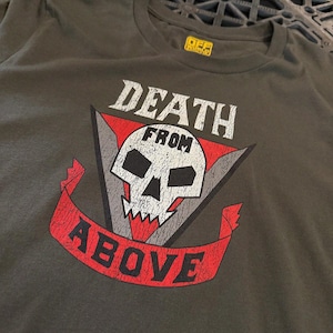 May include: A dark olive green t-shirt featuring a distressed graphic print of a white skull centered on a red and gray triangle. The text "DEATH FROM ABOVE" is printed in white and black lettering, with a yellow brand label inside the collar.