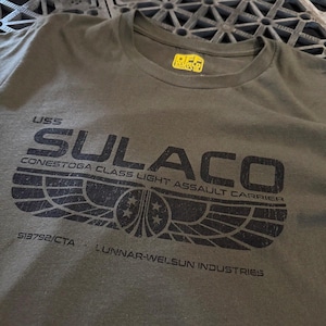 Puede incluir: Una camiseta de cuello redondo color verde oliva con un estampado gráfico negro que dice USS SULACO CONESTOGA CLASS LIGHT ASSAULT CARRIER, 513792/CTA y LUNNAR-WELSUN INDUSTRIES, con un emblema alado y una etiqueta interior amarilla.