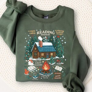 May include: Green sweatshirt with a cozy cabin graphic. The design includes a cabin, trees, a snowman, and text that reads "Reading Retreat" and "For the Cozy Readers". The sweatshirt has long sleeves and a crew neck.