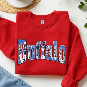 Pode incluir: Um moletom vermelho com a palavra "Buffalo" em azul e branco, apresentando imagens de jogadores de futebol americano. O moletom está dobrado, com um par de jeans azuis visíveis abaixo. Uma xícara de chá e um tapete tecido estão no fundo.