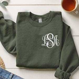 May include: An olive green sweatshirt with a white monogram design. The crew neck sweatshirt is folded on a white wooden surface, with a cup of tea and a pair of jeans in the background.