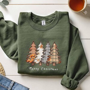 May include: An olive green sweatshirt with a festive design of four Christmas trees in plaid and floral patterns. The words "Merry Christmas" are printed below. A cup of tea and a pair of jeans are in the background.