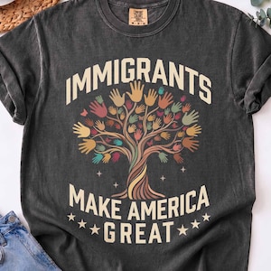 May include: Dark grey t-shirt with the slogan "IMMIGRANTS MAKE AMERICA GREAT" in white. The design shows a tree with colourful handprints as leaves. The shirt is displayed with blue jeans and a cup of coffee.