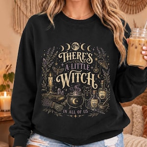 Könnte beinhalten: Ein schwarzes Langarm-Sweatshirt mit einem Grafikdruck und dem Text "THERE'S A LITTLE WITCH IN ALL OF US". Das Design zeigt Illustrationen eines Kessels, einer Katze, einer Kerze, eines Buches sowie botanische Elemente in Gold- und Violetttönen.