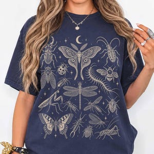 Może przedstawiać: Granatowy t-shirt z krótkim rękawem z kremowym nadrukiem graficznym przedstawiającym różne owady, w tym dużą ćmę, ważkę, motyla, modliszkę i krocionoga, rozmieszczone wśród małych gwiazdek oraz półksiężyca na całej powierzchni koszulki.