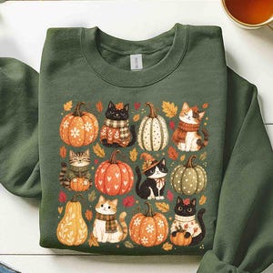 Puede incluir: Una sudadera de cuello redondo en color verde militar con un estampado gráfico de varios gatos con bufandas y sombreros, intercalados con calabazas y hojas de otoño. El diseño incluye gatos naranjas, blancos y negros junto a calabazas estampadas.