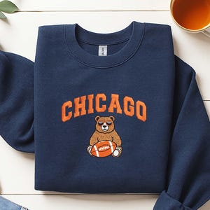 May include: A navy blue crewneck sweatshirt featuring an embroidered graphic of a brown bear wearing sunglasses and holding an American football. The word CHICAGO is embroidered in orange block letters in an arch above the bear graphic.