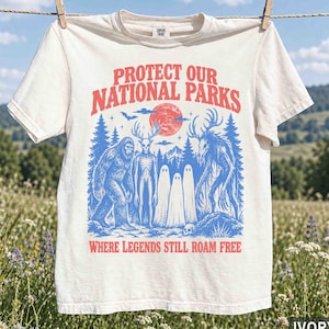 May include: An ivory t-shirt featuring a graphic print with the text PROTECT OUR NATIONAL PARKS and WHERE LEGENDS STILL ROAM FREE. The design includes blue illustrations of Bigfoot, a deer-headed creature, and ghosts against a red moon.