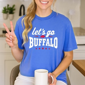 May include: A blue short-sleeved t-shirt featuring the white text "let's go BUFFALO" with a small red buffalo graphic between the words and five red stars underneath. The shirt has a crew neckline and a standard fit, displaying a casual style.