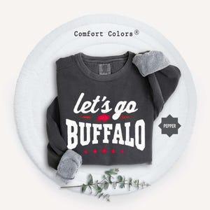 May include: A dark charcoal gray crewneck sweatshirt with the white text "let's go BUFFALO" printed on the front. A small red buffalo graphic is positioned between the words, with four small red stars below. The brand label reads Comfort Colors.