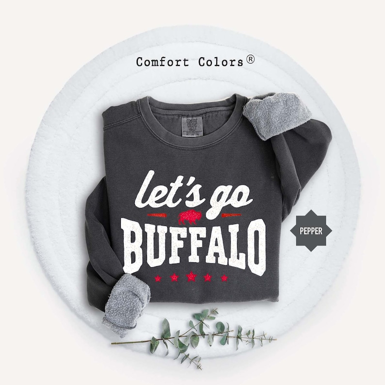 May include: A dark charcoal gray crewneck sweatshirt with the white text "let's go BUFFALO" printed on the front. A small red buffalo graphic is positioned between the words, with four small red stars below. The brand label reads Comfort Colors.
