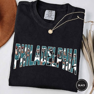 Może przedstawiać: Czarny t-shirt z krótkim rękawem, okrągłym dekoltem i metką marki Comfort Colors. Z przodu znajduje się napis PHILADELPHIA dużymi białymi literami blokowymi, a wewnątrz każdej litery umieszczono grafikę zawodnika futbolu amerykańskiego w turkusowym stroju.