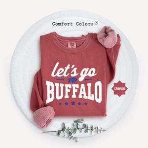 May include: A crimson long-sleeve sweatshirt with a crew neckline. The front features white text that reads let's go BUFFALO, a small blue buffalo graphic, and five blue stars. The Comfort Colors brand label is visible at the inner neck.